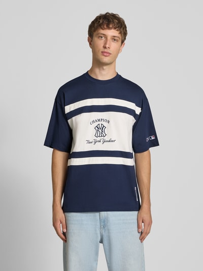 Männermodel trägt Relaxed Fit T-Shirt aus reiner Baumwolle von Champion, blau-weiß mit Logo-Print und Motiv-Stitching, Ganzkörperansicht von vorne. Rückenlänge bei Größe M 70 cm, Rundhalsausschnitt.