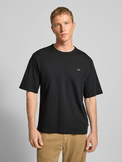 Männermodel trägt T-Shirt mit Label-Print von Levi's® in Schwarz, aus reiner Baumwolle, lockerer Schnitt, gerippter Rundhalsausschnitt, Ganzkörperansicht von vorne.