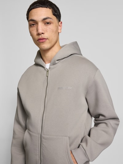 Męski model nosi Sweatjacke mit Känguru-Tasche od REVIEW, bawełniany miks, krój relaxed fit, szary kolor, zamek błyskawiczny, kieszeń typu kangur, długość tyłu w rozmiarze M: 68 cm, długość rękawa w rozmiarze M: 62 cm, zbliżenie na górną część ciała z przodu.