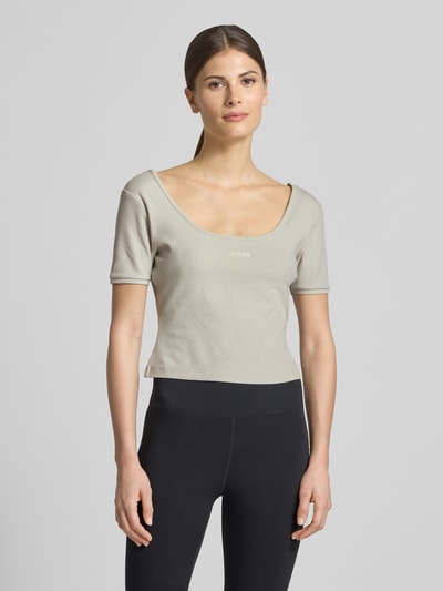 Modelka kobieca nosi Cropped T-Shirt aus Lyocell-Mix Modell 'Circe' od Guess Activewear w kolorze jasnoszarym, logo-stitching, krój slim fit, okrągły dekolt, materiał elastyczny z bawełną, widoczna tekstura, długość tyłu w rozmiarze S: 31 cm, zdjęcie całej sylwetki z przodu.