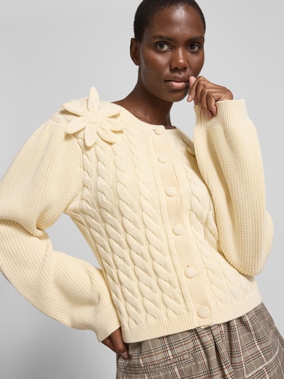 Frauenmodel trägt Strickjacke mit floralem Zierbesatz von BAUM & PFERDGARTEN aus reiner Baumwolle in Creme, taillierte Passform, Rundhalsausschnitt, durchgängige Knopfleiste, Rückenlänge bei Größe S 53 cm, Zopfstrickmuster und grobe Textur; Nahaufnahme Oberkörper.