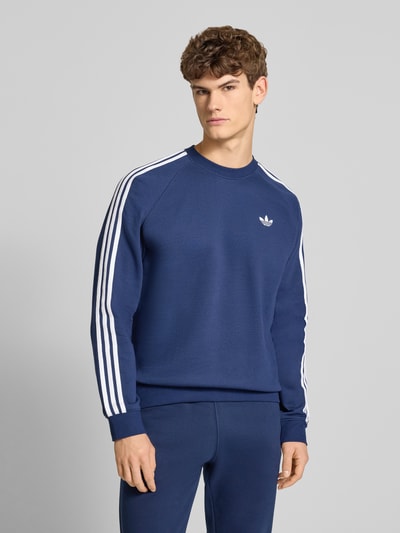 Männermodel trägt Sweatshirt mit Label-Stitching von adidas Originals in Dunkelblau, weiße Streifen, Baumwoll-Mix, Nahaufnahme Oberkörper.