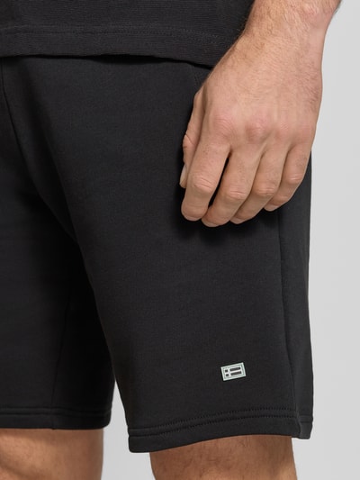 Männermodel trägt Sweatshorts mit Eingrifftaschen von Christian Berg Men in Schwarz, Reine Baumwolle, Regular-Fit, Label-Detail, Nahaufnahme Oberschenkel.