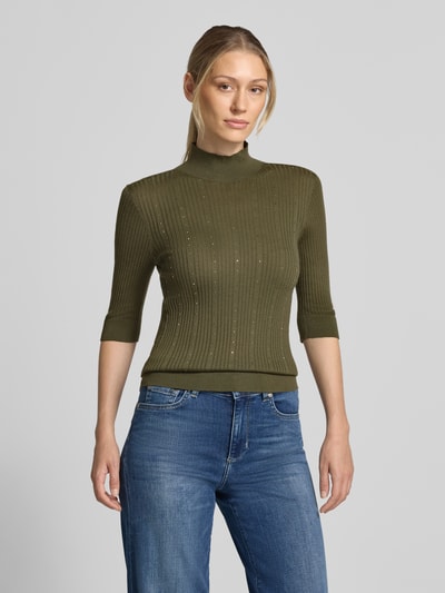 Frauenmodel trägt Slim Fit Longsleeve aus reiner Lanawolle Modell 'Maglia Costine' von Liu Jo White in Olivgrün, semitransparent, feine Rippenstruktur, 1/2-Arm und Stehkragen, kombiniert mit blauer Jeans, Ganzkörperansicht von vorne.