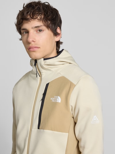 Männermodel trägt Funktionsjacke mit Napoleontasche Modell 'MOUNTAIN ATHLETICS' von The North Face in Beige, Slim-Fit, Kapuze, Nahaufnahme Oberkörper. Rückenlänge 70 cm, Ärmellänge 68 cm, elastische Ärmelabschlüsse, Eingrifftaschen, Reißverschluss.