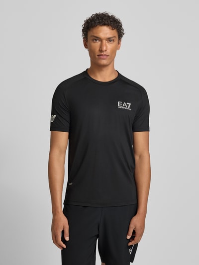 Männermodel trägt T-Shirt mit Label-Print von EA7 Emporio Armani, schwarzer Material-Mix mit Stretch-Anteil, Slim Fit, Ganzkörperansicht von vorne.
