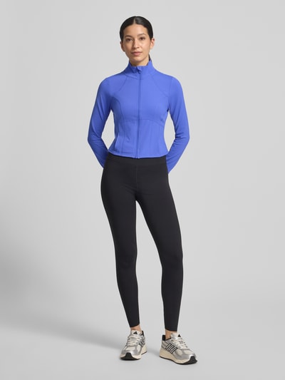 Modelka kobieca nosi Slim Fit Jacke mit Stehkragen Modell 'Storm' od Fabletics, krój slim fit, krótka długość, pętelki na kciuki, kolor niebieski, długość pleców w rozmiarze S: 44 cm, szerokość ramion w rozmiarze S: 36 cm, szerokość klatki piersiowej w rozmiarze S: 42 cm, długość rękawa w rozmiarze S: 74 cm, całopostaciowy widok z przodu.