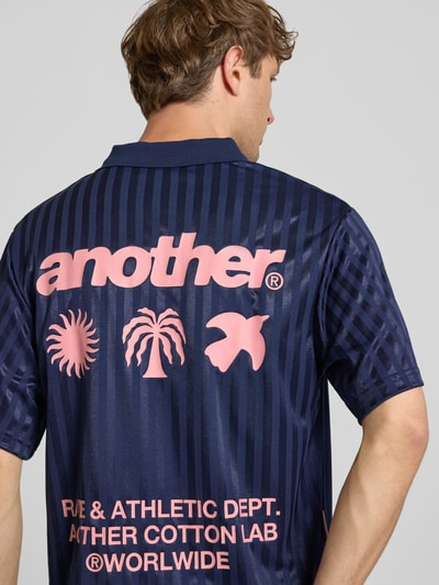 Männermodel trägt Trikot mit Polokragen und Label-Prints von ANOTHER COTTON LAB, krój regular fit, model 'Rave & Athletic Soccer', długość tyłu w rozmiarze M: 70 cm, ciemnoniebieskie paski, różowe nadruki z logo, tkanina o błyszczącej fakturze, Ganzkörperansicht Rückansicht