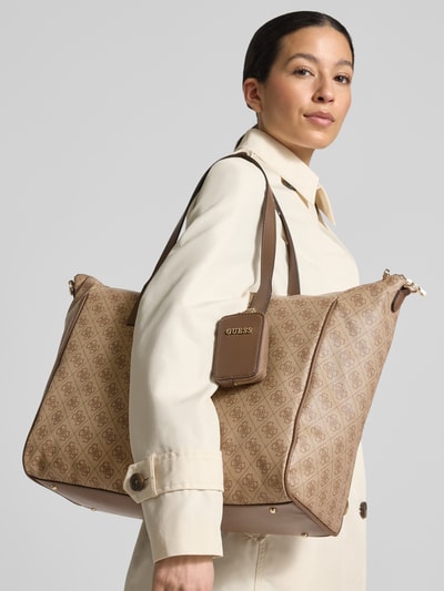 Frauenmodel trägt Shopper mit Logo-Muster Modell 'LATONA CARRYON' von Guess in Beige mit Hauptfach, Tragehenkel und Schulterriemen, Nahaufnahme Oberkörper.