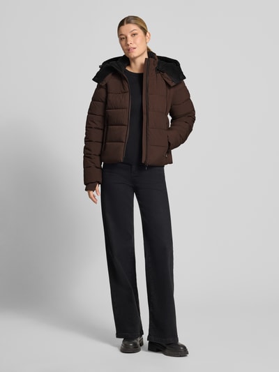 Frauenmodel trägt Jacke mit Daumenloch und Kapuze von NAUTGÅRD in Dunkelbraun, schwarz kombiniert, gerader Schnitt, Ganzkörperansicht von vorne.