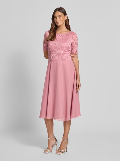 Modelka nosi Knielanges Cocktailkleid mit Spitze und Rundhalsausschnitt von Vera Mont, szyfon, Linia A, długość do kolan, koronka, okrągły dekolt, długość tyłu w rozmiarze 36: 105 cm, szerokość klatki piersiowej w rozmiarze 36: 40 cm, długość rękawów w rozmiarze 36: 28 cm, różowy kolor, faktura koronki i szyfonu, widok całej sylwetki z przodu.