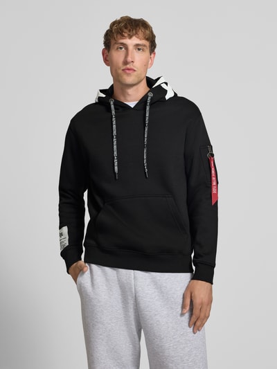 Männermodel trägt Regular Fit Hoodie aus Baumwoll-Mix mit Label-Patch von Alpha Industries in Schwarz, Känguru-Tasche, Ganzkörperansicht von vorne, Rückenlänge 67 cm, Ärmellänge 63 cm, Ärmel-Tasche, Kapuze mit Tunnelzug.
