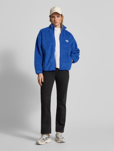 Frauenmodel trägt Jacke aus Teddyfell Modell 'HOKTOWN' von American Vintage in Blau mit Stehkragen, Reißverschluss, Label-Detail, Eingrifftaschen, lockerer Schnitt, Ganzkörperansicht von vorne.