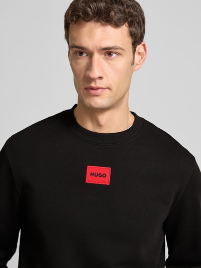 Männermodel trägt Sweatshirt aus Baumwolle Modell 'Diragol' von HUGO in Schwarz mit rotem Label-Patch, Nahaufnahme Oberkörper. Gerader-Schnitt, Gerippter-Rundhalsausschnitt, Gerippte-Abschlüsse, Reine-Baumwolle, Rückenlänge 67 cm, Ärmellänge 64 cm bei Größe M.