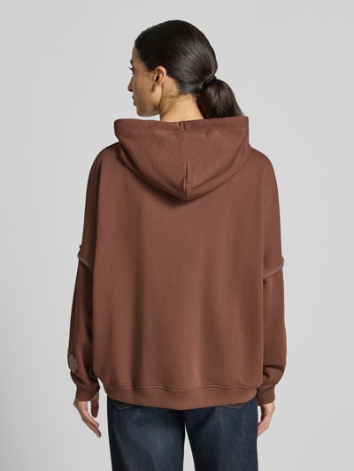 Frauenmodel trägt Oversized Hoodie aus reiner Baumwolle Modell 'L(I)EBE' von miss goodlife in Braun mit gerippten Abschlüssen und Känguru-Tasche, Detailverzierung mit Pailletten und Strasssteinen, Ganzkörperansicht Rückansicht.