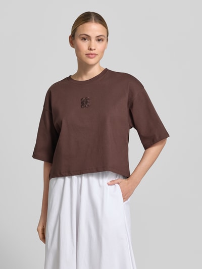 Frauenmodel trägt Cropped Boxy Fit T-Shirt mit Rundhalsausschnitt von Smith and Soul, reine Baumwolle, dunkelbraun mit dezentem Stitching, Rückseite länger bei Größe S: 49 cm, glatte Textur, Ganzkörperansicht von vorne.