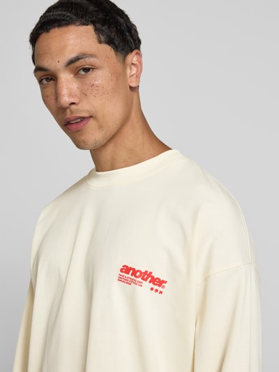 Męski model nosi bluzkę z długim rękawem z nadrukiem z logo i okrągłym dekoltem od ANOTHER COTTON LAB, bawełna, krój relaxed fit, długość tyłu w rozmiarze M: 65 cm, jasny kolor, gładka tekstura, zbliżenie na górną część ciała