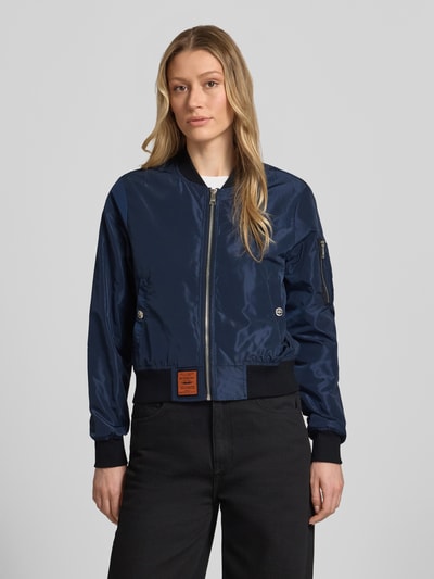 Frauenmodel trägt Bomberjacke mit Label-Patch und Reißverschlusstasche am Ärmel von Original Bombers in Dunkelblau mit Stehkragen, Logo-Stitching, Druckknopfverschluss an seitlichen Eingrifftaschen, elastische Abschlüsse, Rückenlänge 53 cm, Schulterbreite 37 cm, Brustweite 48 cm, Ärmellänge 63 cm, Lockerer-Schnitt, glatte Textur, Ganzkörperansicht von vorne.