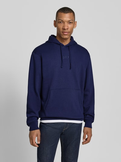 Männermodel trägt Oversized Hoodie mit Logo-Stitching von GAP in Dunkelblau, mit Kapuze, Känguru-Tasche, Baumwoll-Mix, Ganzkörperansicht von vorne.