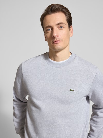 Męski model nosi Regular Fit Sweatshirt aus Baumwoll-Mix von Lacoste, efekt: jednolity kolor, naszywka z logo, okrągły dekolt, długi rękaw, prążkowane wykończenia, mieszanka bawełny, dzianina dresowa, kolor jasnoszary, zbliżenie na górną część ciała z przodu.