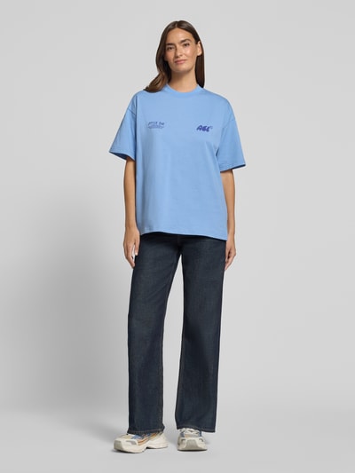 Modelka nosi damski t-shirt z bawełny o kroju relaxed fit, model 'Oyster Bar' z nadrukiem z logo ANOTHER COTTON LAB, okrągły dekolt, długość tyłu 61 cm w rozmiarze S, niebieski kolor, widok całej sylwetki z przodu.