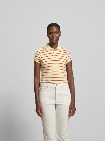 Frauenmodel trägt Cropped Poloshirt aus reiner Baumwolle von Rabanne mit Polokragen, verlängerte Rückseite, kurzärmeligem Design, Rückenlänge bei Größe S 47 cm, gestreiftes Muster in Gelb und Rot auf glatter Textur, Ganzkörperansicht von vorne.