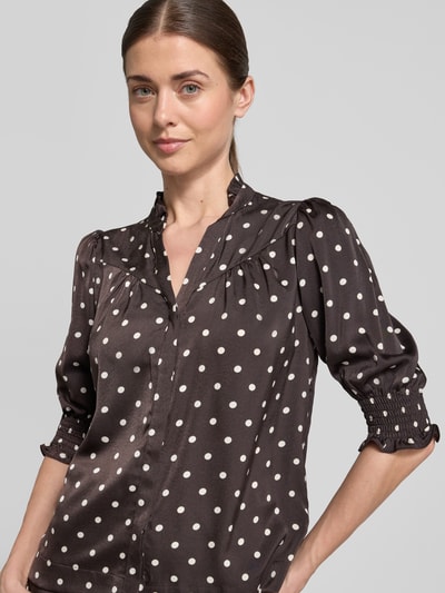 Kobiece modelka nosi Regular Fit Bluzę ze stójką model 'Diana' od Neo Noir, czarna w białe grochy, obszycie falbanami, rękaw o dł. 1/2, elastyczne wykończenia, długość tyłu w rozmiarze 36: 59 cm, długość rękawów: 41 cm, zbliżenie na górną część ciała, widok z przodu.
