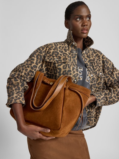 Frauenmodel trägt Tote Bag mit Label-Detail von Tory Burch in Braun mit Zugband, Hauptfach, Lange Tragehenkel, Innenfach, Ganzkörperansicht von vorne.