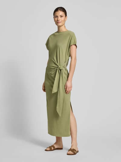 Frauenmodel trägt Midikleid aus reiner Baumwolle von Scotch & Soda in Khaki mit Knoten-Detail und Wickel-Optik, taillierte Passform, Ganzkörperansicht von vorne.
