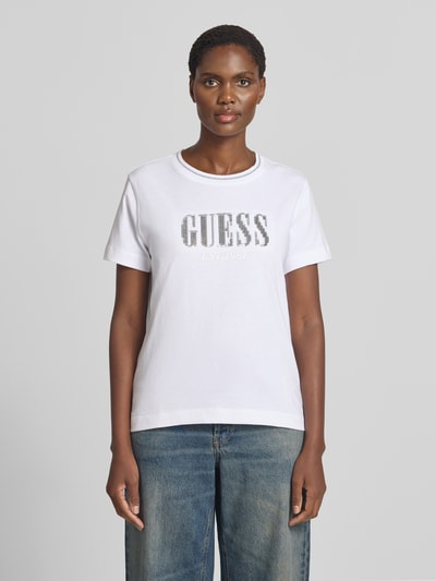 Vrouwenmodel draagt t-shirt uit puur katoen met logodetail van GUESS, getailleerde pasvorm, met pailletten, ronde hals, ruglengte maat S 58 cm. Ganzkörperansicht von vorne.
