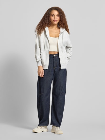 Modelka nosi Oversized Fit Sweatjacke mit geteilter Kängurutasche od Gina Tricot, mieszanka bawełny, krój oversized fit, kieszeń kangurka, kaptur z tunelem, elastyczne wykończenia, jasnoszara bluza, ciemnogranatowe spodnie, widoczne tekstury tkanin, całkowity widok z przodu.