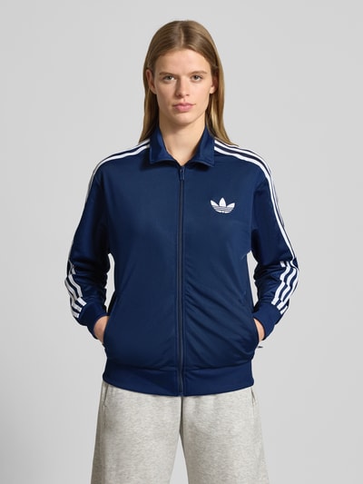Vrouwenmodel draagt Sweatjacke mit Stehkragen van adidas Originals in donkerblauw met witte strepen, losse pasvorm, ribboorden, Ganzkörperansicht von vorne.
