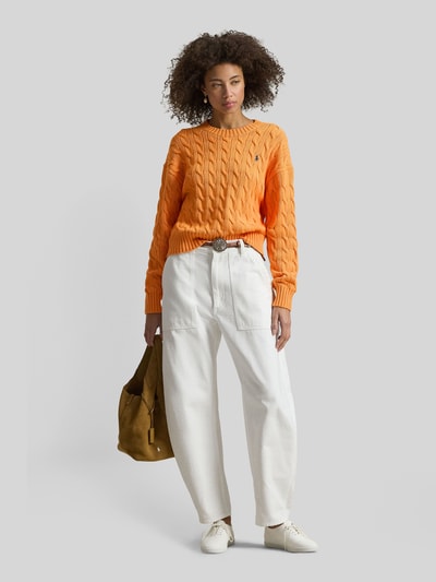 Vrouwenmodel draagt Regular Fit Strickpullover aus reiner Baumwolle van Polo Ralph Lauren in effen oranje met kabelpatroon en logo-stiksel, lange mouw, geribde boorden, grof gebreide pure katoen, voorkant volledig zichtbaar, geheellichaamsaanzicht van voren.