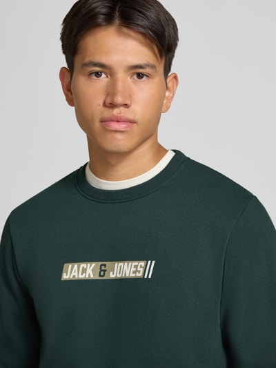 Männermodel trägt Regular Fit Sweatshirt mit Baumwoll-Anteil Modell 'Munk' von JACK & JONES in Dunkelgrün, Nahaufnahme Oberkörper. Material-Mix, Logo-Print, Rundhalsausschnitt, elastische Abschlüsse.