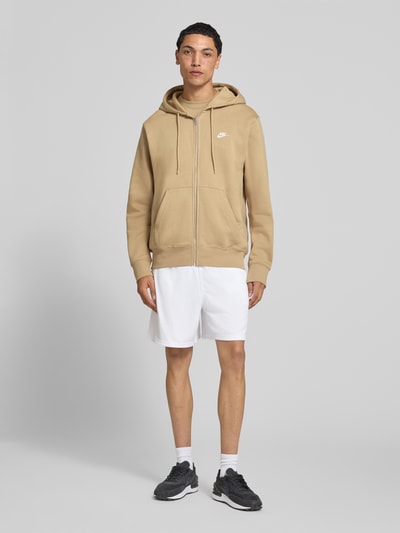 Männermodel trägt Regular Fit Sweatjacke aus Baumwoll-Mix von Nike in Beige mit Frontreißverschluss, kombiniert mit weißen Shorts, Ganzkörperansicht von vorne. Herren Sweatjacke von Nike.