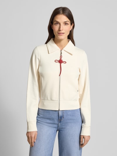 Modelka damska nosi Slim Fit Sweatjacke aus reiner Baumwolle Modell 'EROICO' von MAX&Co., kremowy kolor, gładka faktura, detal czerwona kokardka przy zamku, długi rękaw, kołnierz koszulowy, metaliczne akcenty, klasyczne ściągacze, boczne kieszenie, widok całej sylwetki z przodu.