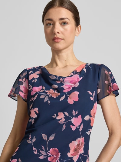 Frauenmodel trägt Knielanges Cocktailkleid mit Wasserfall-Ausschnitt von Christian Berg Cocktail aus Chiffon mit Schmetterlingsärmel, taillierter Schnitt, florales Muster, Ganzkörperansicht von vorne.
