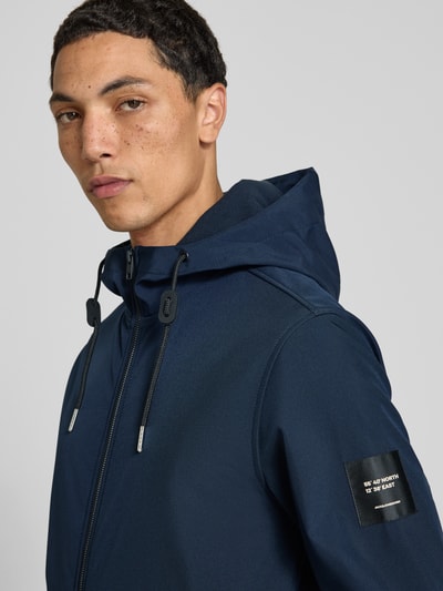 Männermodel trägt Regular Fit Jacke mit Logo-Detail von JACK & JONES in Dunkelblau, glatte Textur, Nahaufnahme Oberkörper. Softshell, Kapuze mit Tunnelzug, Eingrifftaschen mit Reißverschluss, elastische Abschlüsse, Rückenlänge 66 cm, Schulterbreite 46 cm, Brustweite 53 cm, Ärmellänge 67 cm.