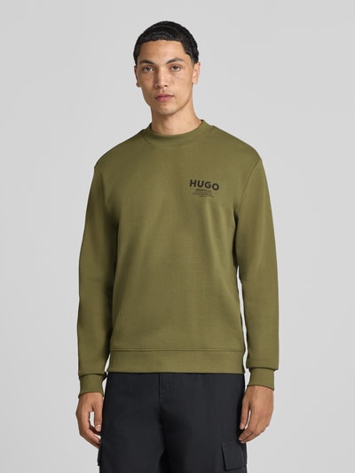 Męski model nosi Regular Fit Sweatshirt aus reiner Baumwolle model 'Nespon' od HUGO, oliwkowa z czarnym nadrukiem, bluza bawełniana, widok całej sylwetki z przodu, kolekcja HUGO Blue.