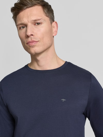 Mężczyzna model nosi bluzkę z długim rękawem z okrągłym dekoltem i logo Longsleeve mit Rundhalsausschnitt und Logo von Fynch-Hatton, wykonana z czystej bawełny, krój regular fit, jednokolorowy, długość tyłu 70 cm w rozmiarze M, kolor granatowy, detal logo na piersi, jednolita faktura, zbliżenie na górną część ciała.