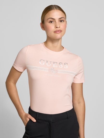 Frauenmodel trägt T-Shirt mit Strasssteinbesatz Modell 'SIENA' von Guess, zartrosa Farbton, Logo-Print auf der Front, Nahaufnahme Oberkörper. Damen T-Shirt aus Baumwoll-Mix mit Stretch-Anteil, gerader Schnitt, Rundhalsausschnitt.