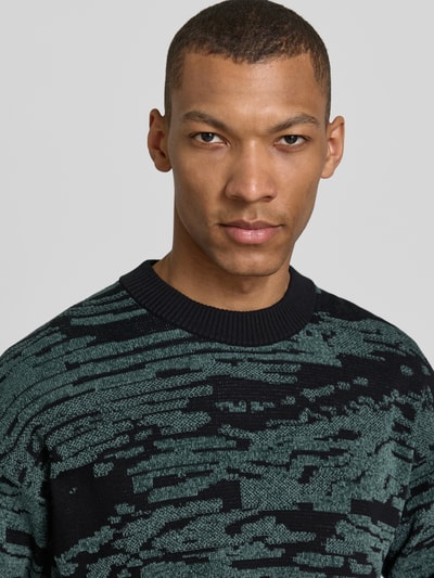 Mężczyzna nosi Regular Fit Strickpullover mit grafischem Jacquard-Muster Modell 'Andscape' od BOSS Orange, bawełniany mix, ciemny zielony i czarny, wzór allover, okrągły dekolt, długi rękaw, widok na górną część ciała z przodu.