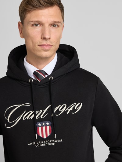 Männermodel trägt Hoodie mit Label-Stitching und Känguru-Tasche von Gant, schwarzer Baumwoll-Mix, Regular-Fit, Nahaufnahme Oberkörper.