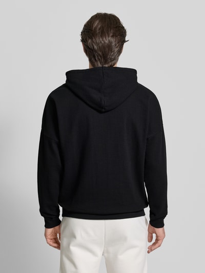 Męski model nosi Relaxed Fit Hoodie aus Baumwoll-Mix od HUGO, efekt: jednolity kolor, wyhaftowane logo, faktura wafla, czarny, długość tyłu 71 cm, szerokość klatki piersiowej 60 cm, długi rękaw, prążkowane zakończenia, mieszanka bawełny, Ganzkörperansicht Rückansicht.