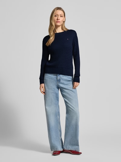Frauenmodel trägt Slim Fit Pullover aus reiner Baumwolle von Tommy Hilfiger in Dunkelblau, Optik: Unifarben, Strickoptik mit Zopfmuster, Logo-Detail, Rundhals, Langarm, Ärmelabschluss gerippt, Rumpfabschluss gerippt, Materialverarbeitung: Strick, Ganzkörperansicht von vorne.