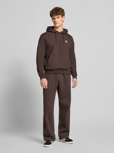 Męski model nosi Hoodie mit Kapuze od adidas Originals z bawełnianej mieszanki, luźny krój, kaptur, tunel, naszywka z logo, kieszeń kangurka, ściągacze, długość tyłu i rękawa 66 cm w rozmiarze M, brązowy kolor, jednolita faktura, pełny widok z przodu