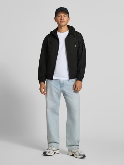 Männermodel trägt Regular Fit Jacke mit Logo-Detail von JACK & JONES, schwarz, Softshell, Kapuze, Reißverschlusstaschen, Ganzkörperansicht von vorne