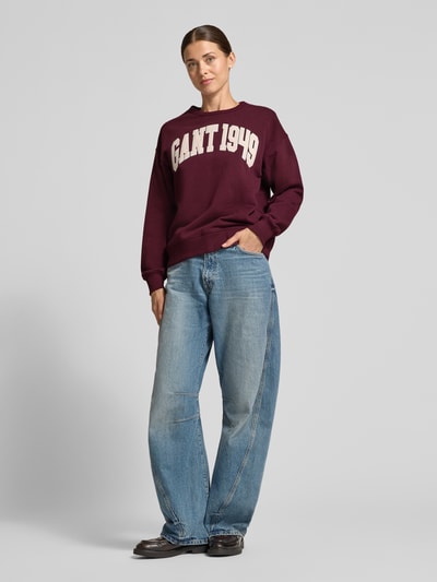 Frauenmodel trägt Relaxed Fit Sweatshirt aus reiner Baumwolle von GANT in Dunkelrot mit weißem Schriftzug, kombiniert mit blauer Jeans, Ganzkörperansicht von vorne. Damen Sweatshirt von GANT.