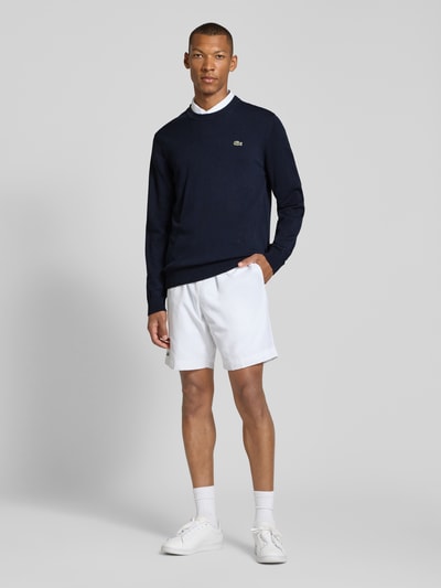Männermodel trägt Regular Fit Pullover aus Baumwoll-Mix von Lacoste in Dunkelblau, kombiniert mit weißen Shorts, Ganzkörperansicht von vorne. Herren Pullover von Lacoste