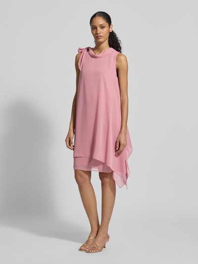Modelka kobieca nosi Relaxed Fit Kleid mit Stehkragen od Vera Mont, półprzezroczysty, asymetryczny różowy materiał, ozdobna kokarda, podszewka kryjąca, krój relaxed fit, widok całej sylwetki z przodu.